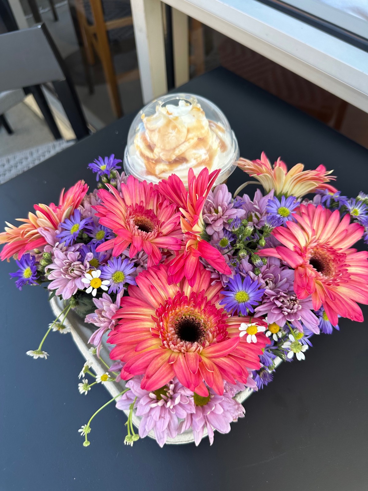 Vibrant pink gerbera daisies and purple flowers surround a coffee